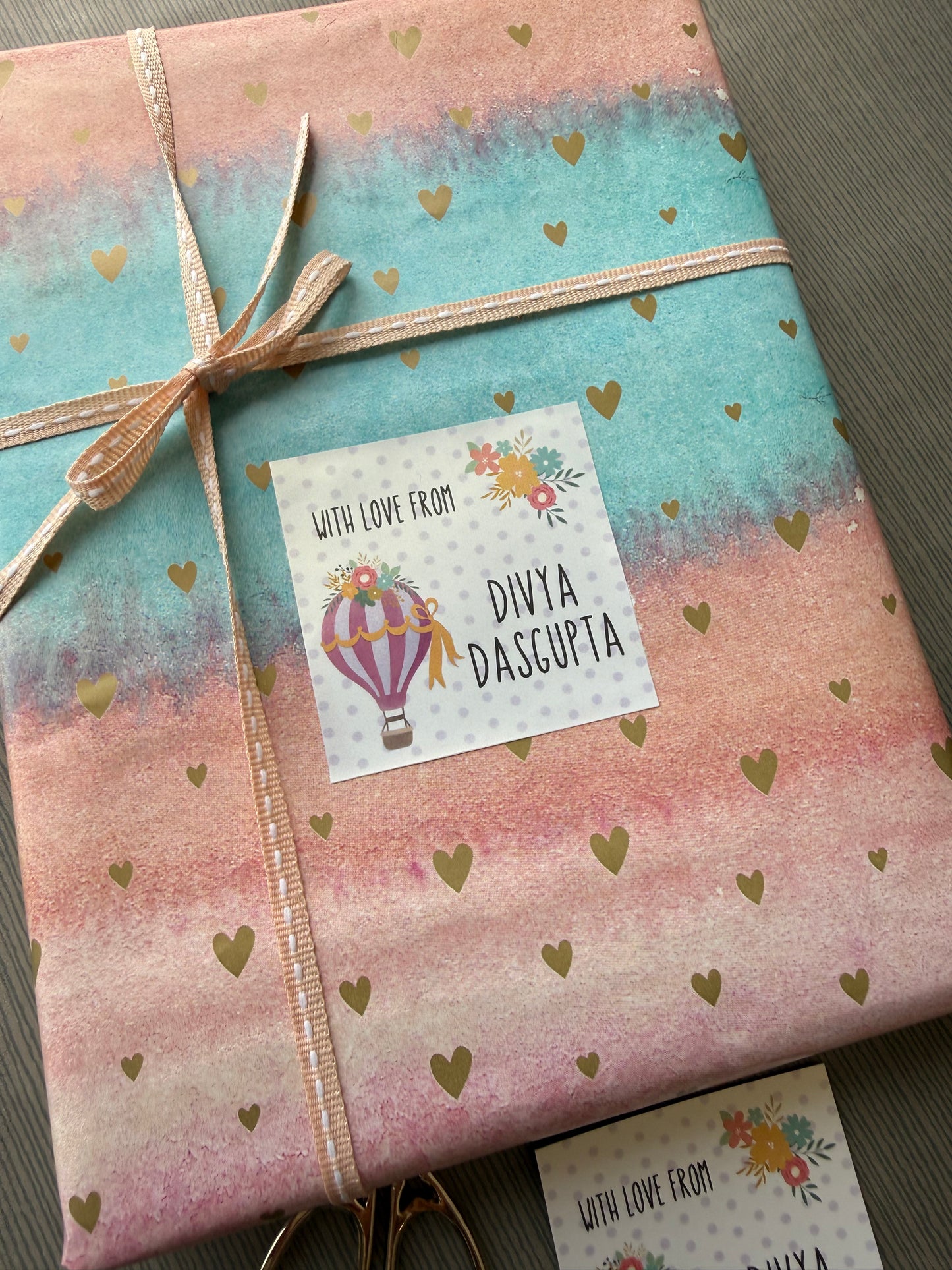 A wrapped gift box with pastel pink and turquoise watercolor-style paper, scattered gold heart motifs, and a peach ribbon tied in a bow. A square printed card is attached to the front, and a matching card is partially visible at the bottom edge.