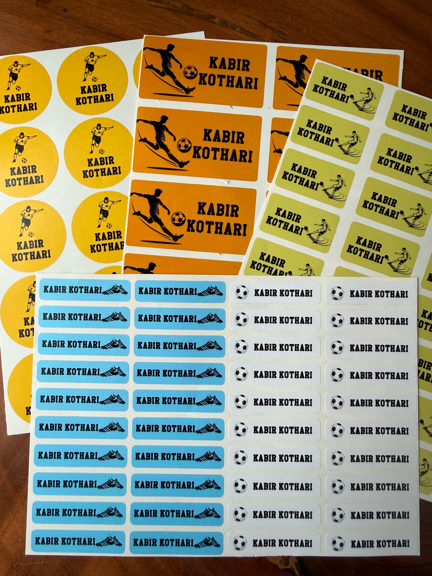 Several sheets of waterproof name label round and rectangular stickers  yellow, orange, and light blue backgrounds, with black silhouettes of football  players and soccer balls.