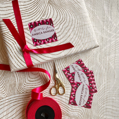 A wrapped gift box tied with a red ribbon, shown with matching red ribbon spool, gold-handled scissors, and several decorative gift tags printed with the Ambica Ranger logo on a light wood-grain surface.