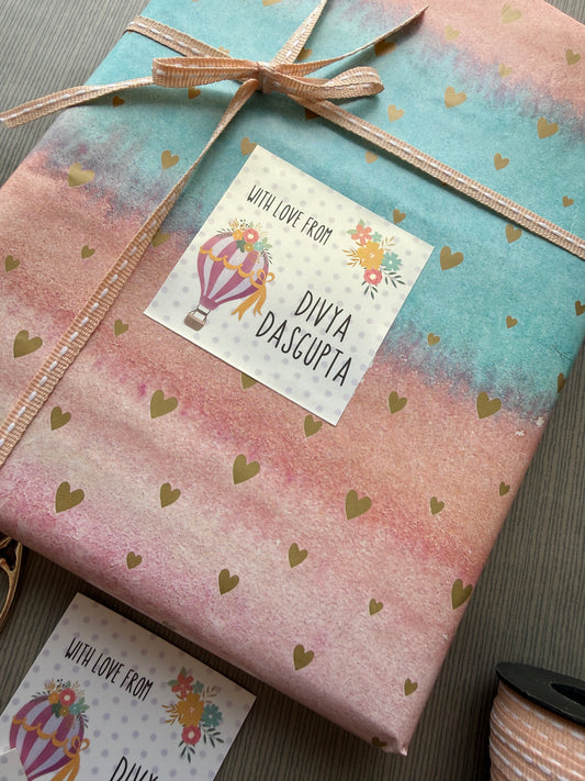A gift-wrapped package on a dark wood surface, featuring pastel watercolor paper with a blue-green left side, pink right side, scattered gold hearts, a beige ribbon tied in a bow, and a white printed card that reads 'LIFE LOVE FROM DIVIA DASCOPIA' with a hot air balloon illustration.