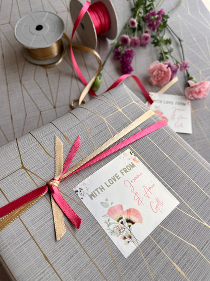 A wrapped gift box with a gray and gold geometric patterned paper, tied with a pink and gold ribbon, and adorned with a decorative card. Pink and gold ribbon spools and pink flowers are arranged on a matching patterned surface.