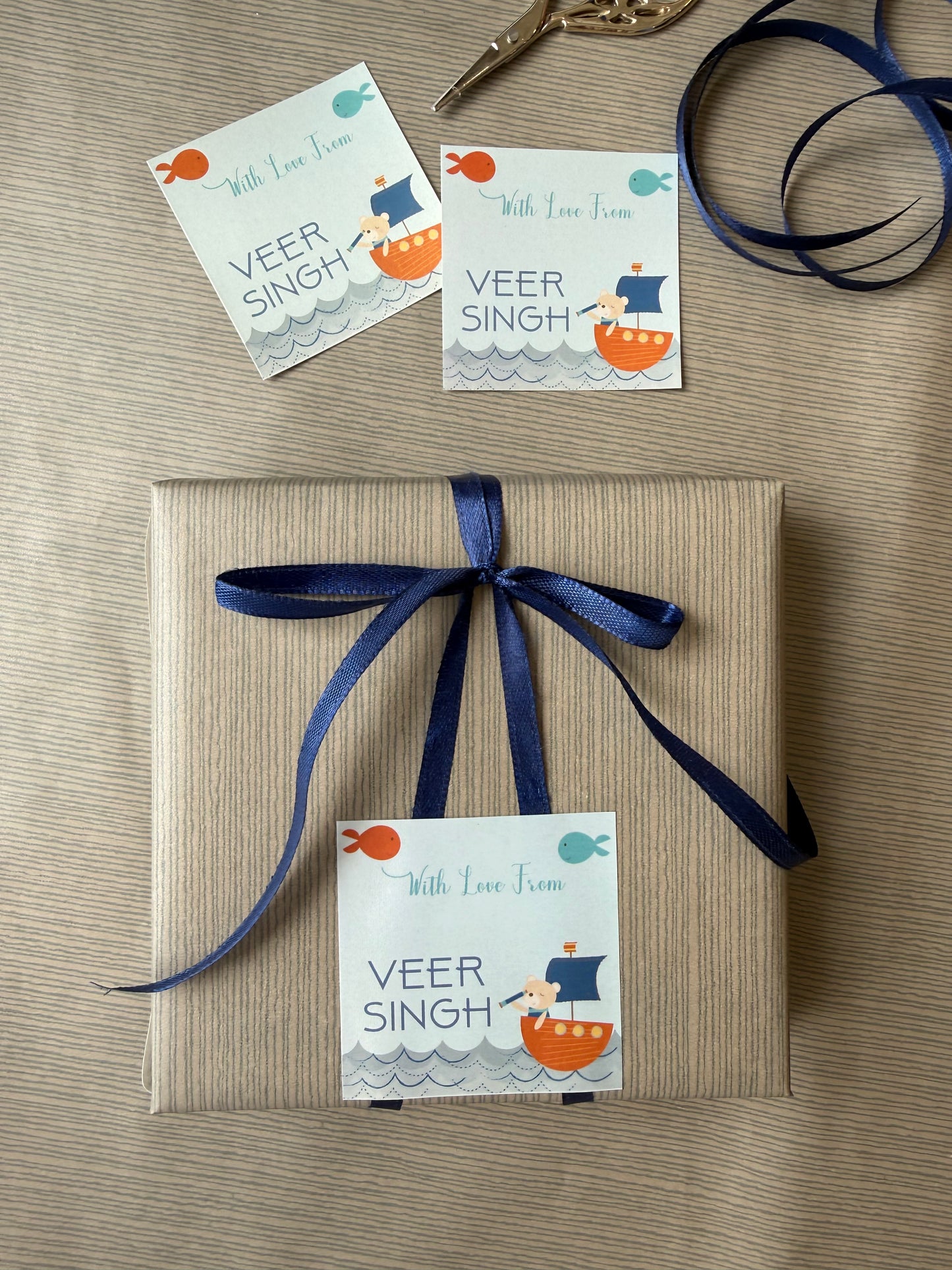 A wrapped gift box tied with a navy ribbon and a printed 'With Love From VEER SINGH' label, shown on a beige textured surface with two matching labels and ribbon in the background.