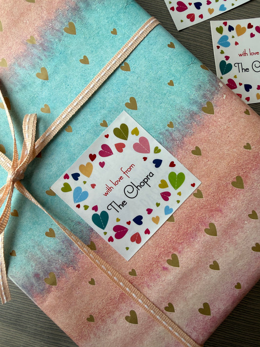 A gift-wrapped package tied with a beige ribbon, featuring a pastel watercolor design with gold heart patterns. A white square sticker on the front reads 'The Shop' with colorful hearts and the phrase 'with love from'.