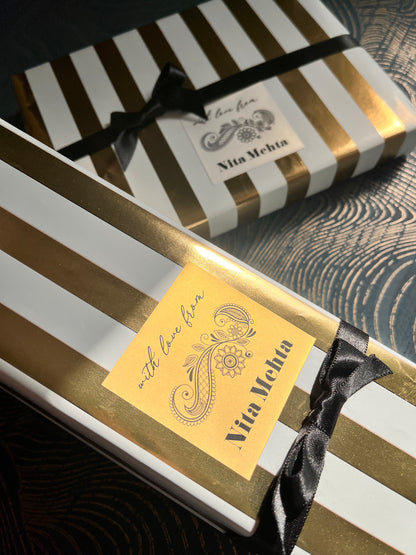Two elegant gift boxes with white and gold vertical stripes, tied with black satin ribbons and adorned with gold labels. One label reads 'Via Meeta' and 'with Love from'.