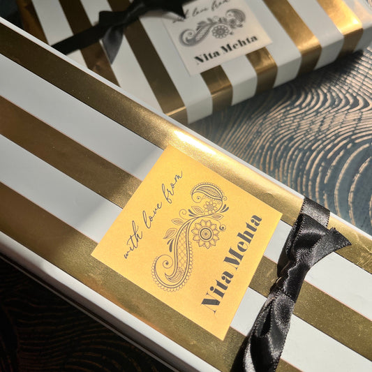 Two elegant gift boxes with white and gold vertical stripes, tied with black satin ribbons and adorned with gold labels. One label reads 'Via Meeta' and 'with Love from'.