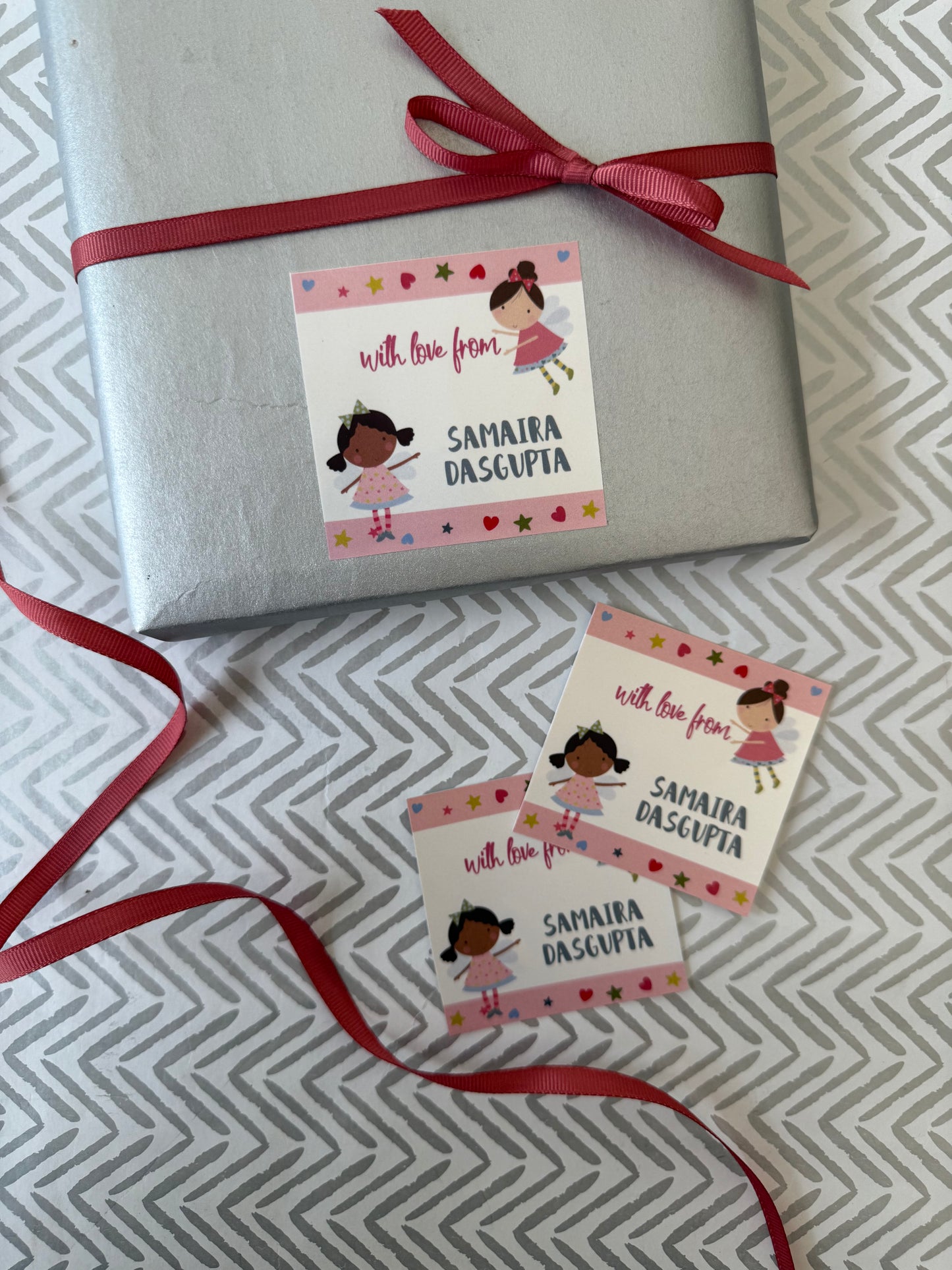 A silver gift box tied with a red ribbon and two matching personalized gift tags with cartoon girl illustrations and the text 'with love from SAMAIRA DASCUPIA' on a gray patterned surface.
