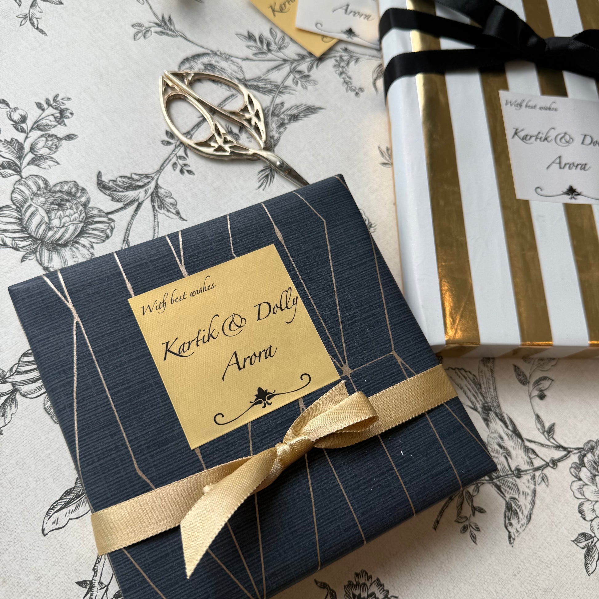 A wrapped gift box with a navy blue textured cover, gold geometric line details, and a gold ribbon bow, placed on a floral tablecloth next to scissors and another wrapped package.