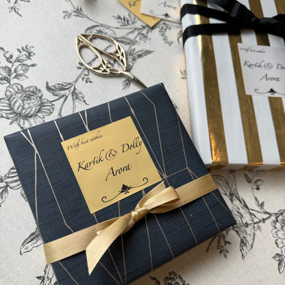 A wrapped gift box with a navy blue textured cover, gold geometric line details, and a gold ribbon bow, placed on a floral tablecloth next to scissors and another wrapped package.