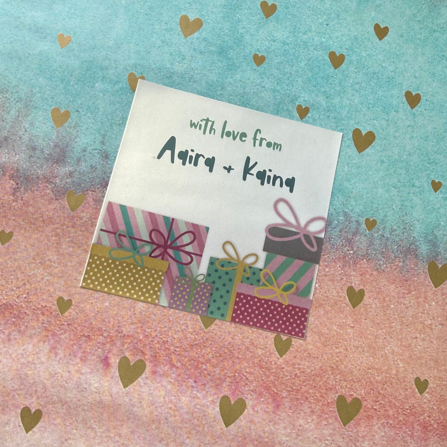 A wrapped gift box or package covered in pastel watercolor paper with gold heart patterns. The left side is light blue, the right side is light pink, and the center features a vertical gradient blending from blue to pink.