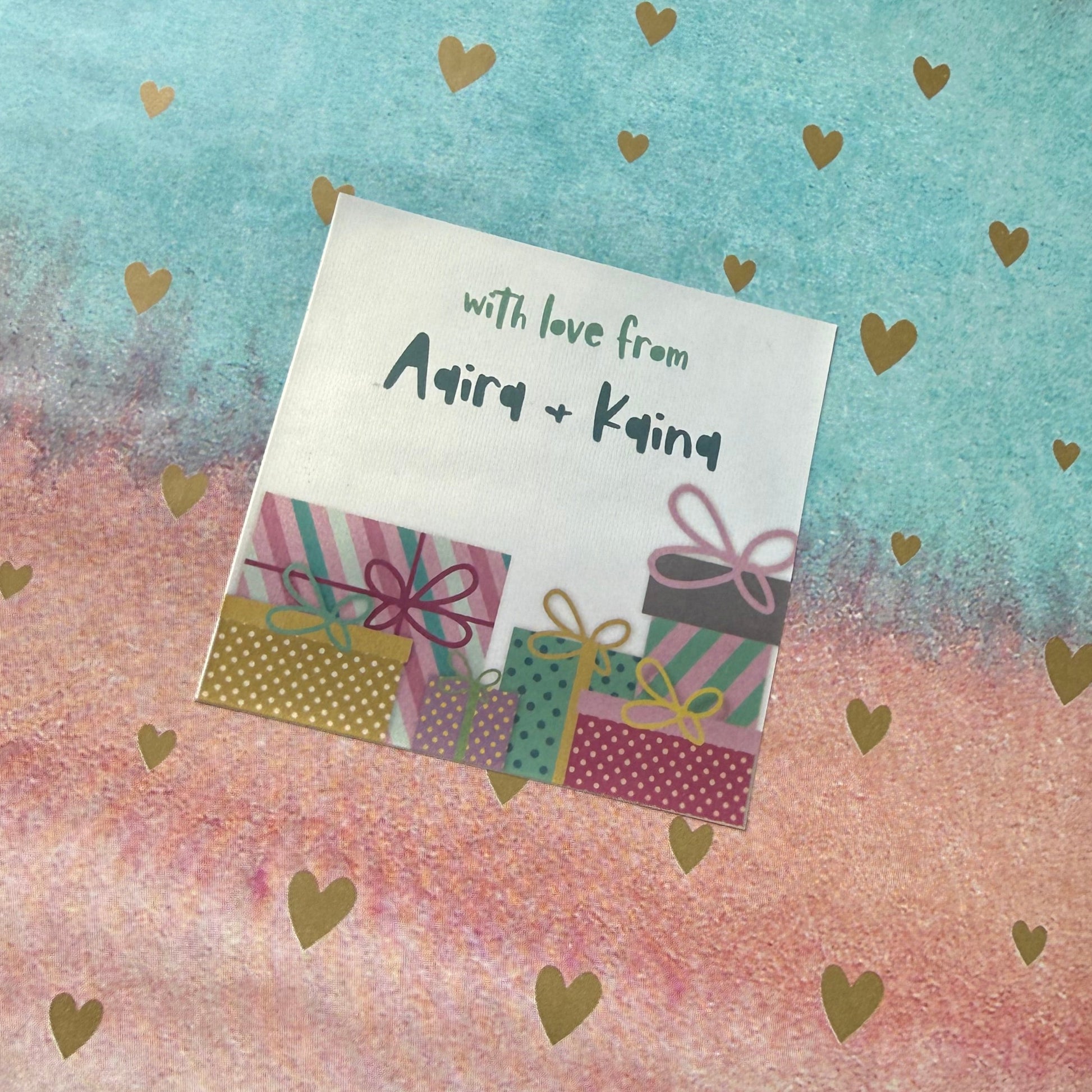 A wrapped gift box or package covered in pastel watercolor paper with gold heart patterns. The left side is light blue, the right side is light pink, and the center features a vertical gradient blending from blue to pink.