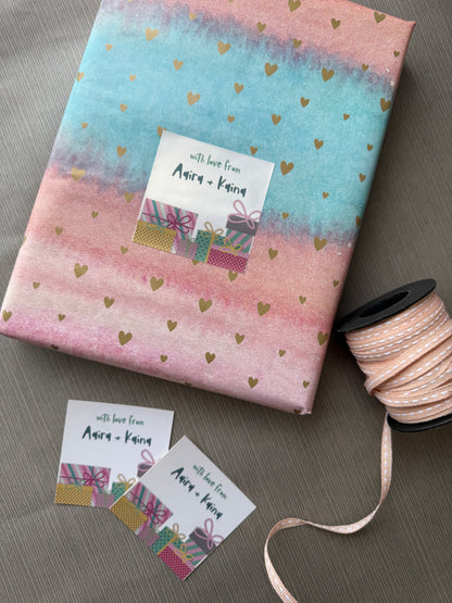 A wrapped gift box with pastel pink and blue watercolor-style paper, scattered gold heart confetti, and a white gift tag reading 'with love from Anna + Karen.' A spool of matching pink ribbon and two small matching tags are arranged on a gray surface.