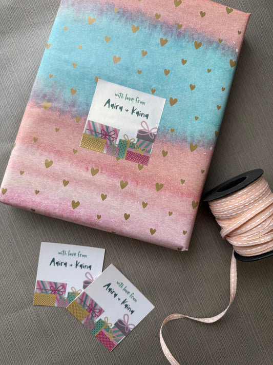 A wrapped gift box with pastel pink and blue watercolor-style paper, scattered gold heart confetti, and a white gift tag reading 'with love from Anna + Karen.' A spool of matching pink ribbon and two small matching tags are arranged on a gray surface.