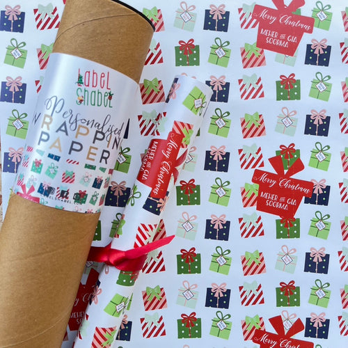 A roll of personalized wrapping paper with Christmas-themed patterns, gifts, and text, placed on a surface with Christmas decorations.