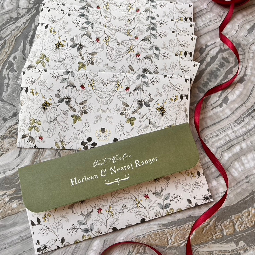 A stack of personalized floral gift envelopes with a green flap reading 'Best Wishes Harleen & Neeraj Ranger,' arranged on a gray marble surface with a red ribbon.