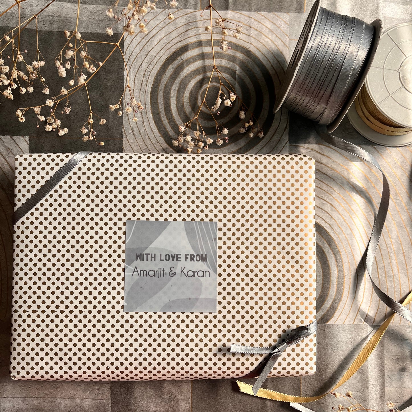 A square gift box wrapped in beige paper with a dense black polka dot pattern, featuring a central  gift label that reads 'WITH LOVE FROM Amira & Karam' and a black ribbon tied around it. The box is styled on a gray wood-patterned surface alongside a spool of gray ribbon and dried baby's breath flowers.