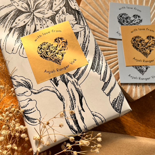 A wrapped gift box with black floral line art on cream paper, adorned with a square gold foil label reading 'with love from Anjali Ranger Virk' and a decorative heart motif. Two matching cards are placed nearby on a wooden surface with dried flowers.