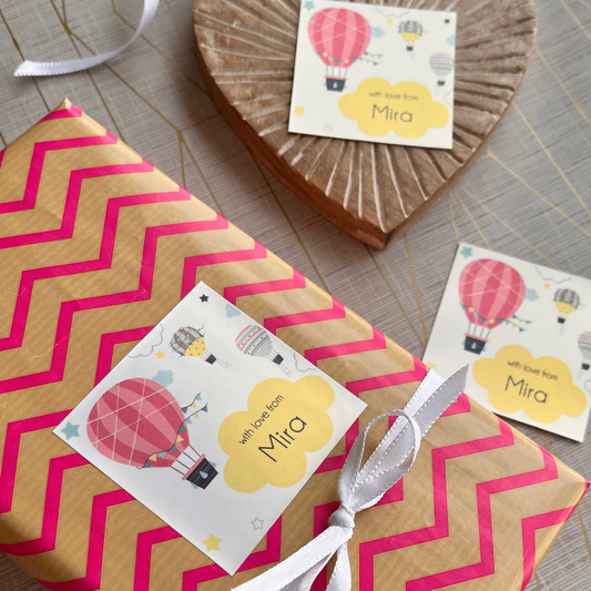 A wrapped gift box with a pink and beige chevron pattern, tied with a white ribbon and decorated with a custom hot air balloon tag reading 'Mira'. A wooden heart ornament and another similar tag are displayed on a neutral textured background.