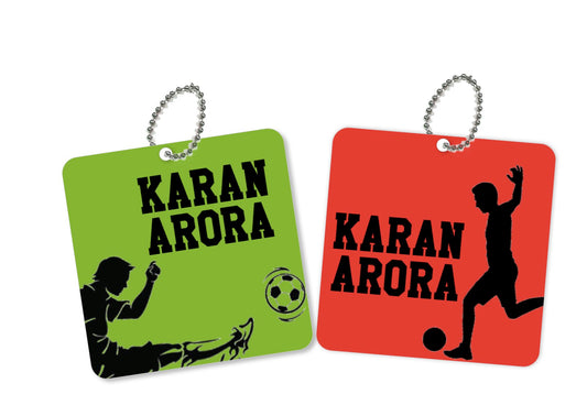 Two square metal bag tags with a soccer player silhouette and the name 'Karan Arora' written on them. One is green with a black and white design, and the other is red with a black and white design.