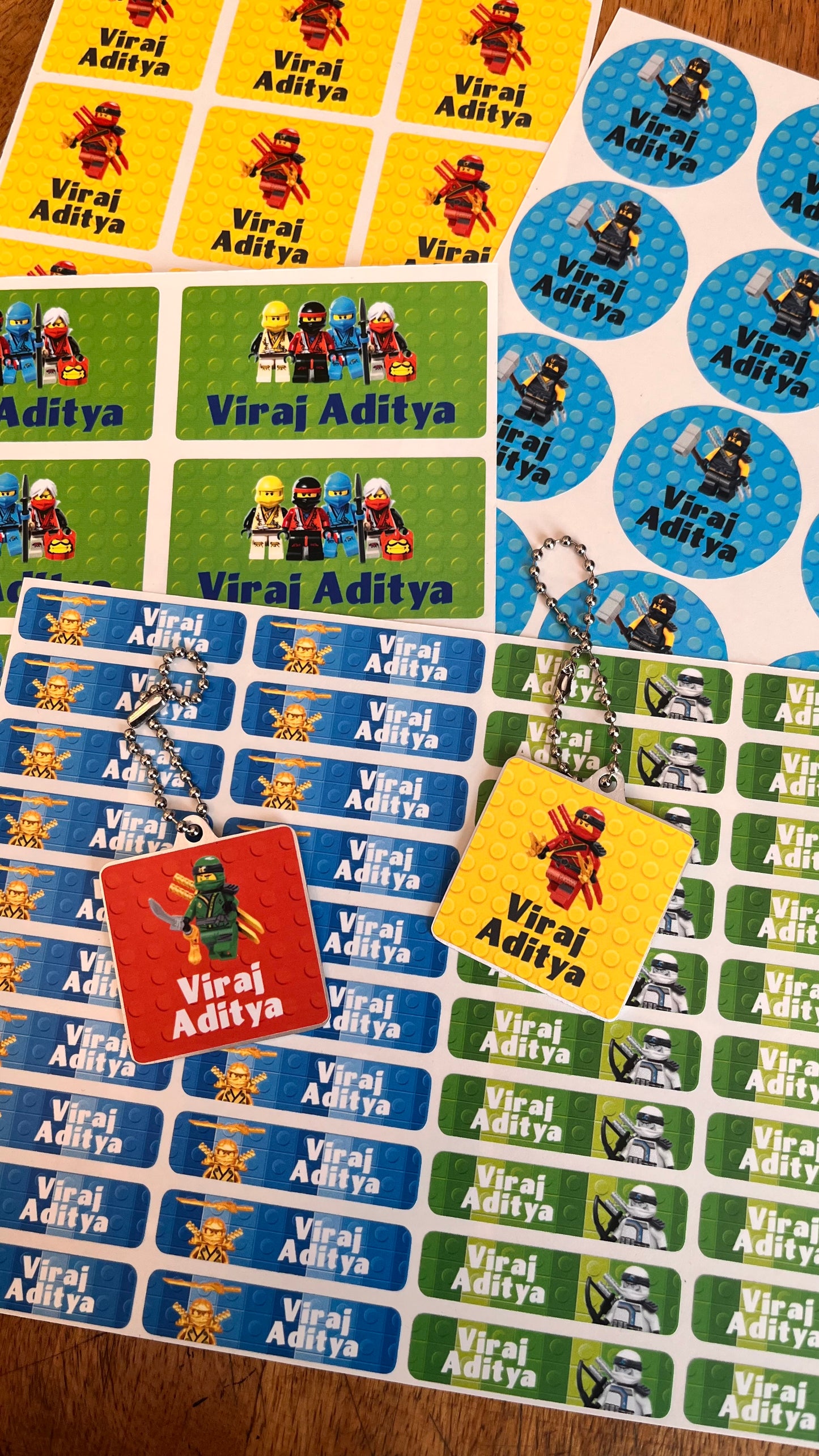 Custom printed sticker sheets and keychains featuring Lego minifigures and the name 'Viraj Aditya' on a wooden surface.