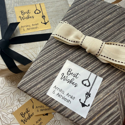 A wrapped gift box with a wood-grain patterned paper wrap, a cream ribbon with black stitching, and a white 'Best Wishes' card with an anchor illustration. A black ribbon bow is tied on the left, and two small kraft paper cards are visible on a patterned surface.