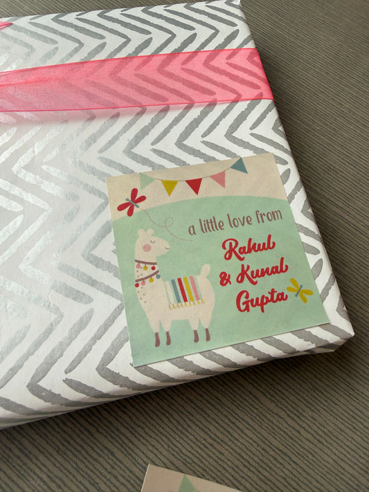 A wrapped gift box with gray and white chevron-patterned paper, a pink ribbon, and a personalized card featuring a llama illustration and the text 'a little love from Rahul & Kunal Gupta'.