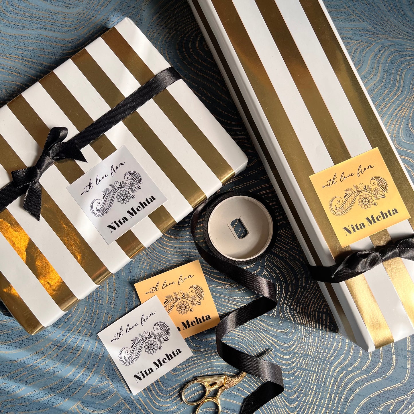 Three gift boxes wrapped in white and metallic gold striped paper, tied with black ribbons, arranged on a blue textured fabric background. 