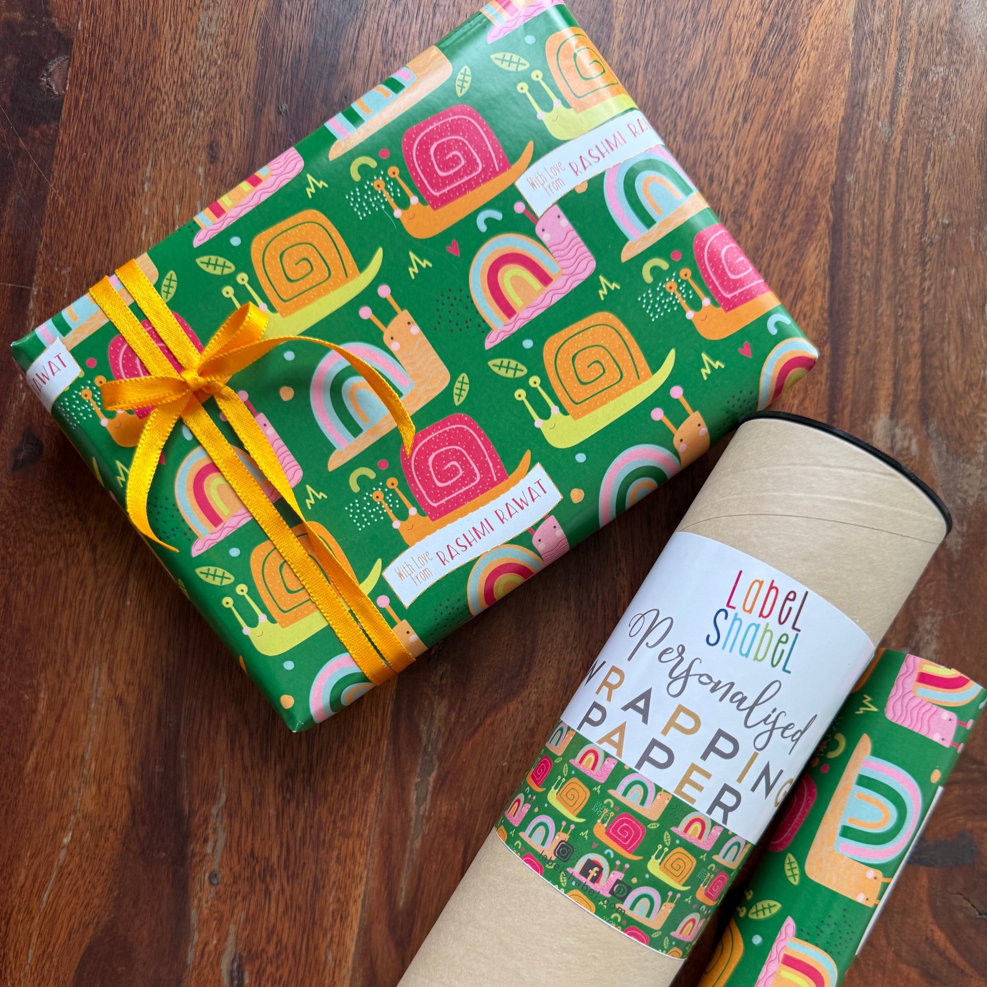 A roll of colorful patterned wrapping paper and a wrapped gift box on a dark wooden surface. The wrapping paper has a green background with pink, orange, and yellow retro  snail shapes and a white label that reads personalised wrapping paper