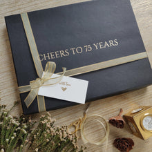 a black box with gold lettering stating cheers to 75 years tied in a golden ribbon with a gift tag saying with love
