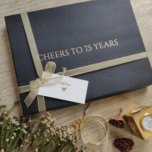 a black box with gold lettering stating cheers to 75 years tied in a golden ribbon with a gift tag saying with love