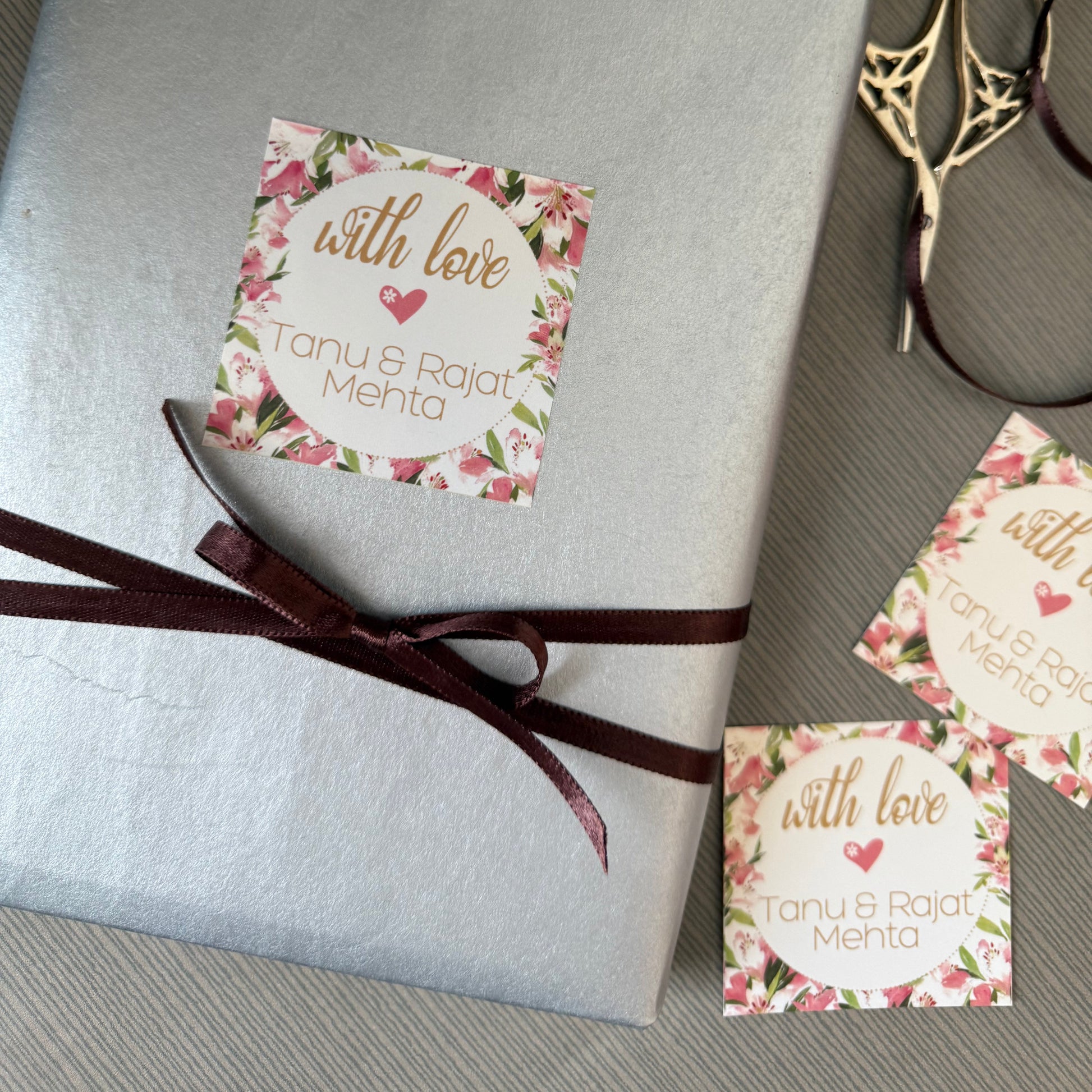 A wrapped gift box in textured silver paper with a dark brown ribbon and a floral 'with love Tanu & Rajat Mehta' sticker, styled on a gray textured surface with gold scissors and two additional stickers.