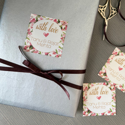 A wrapped gift box in textured silver paper with a dark brown ribbon and a floral 'with love Tanu & Rajat Mehta' sticker, styled on a gray textured surface with gold scissors and two additional stickers.