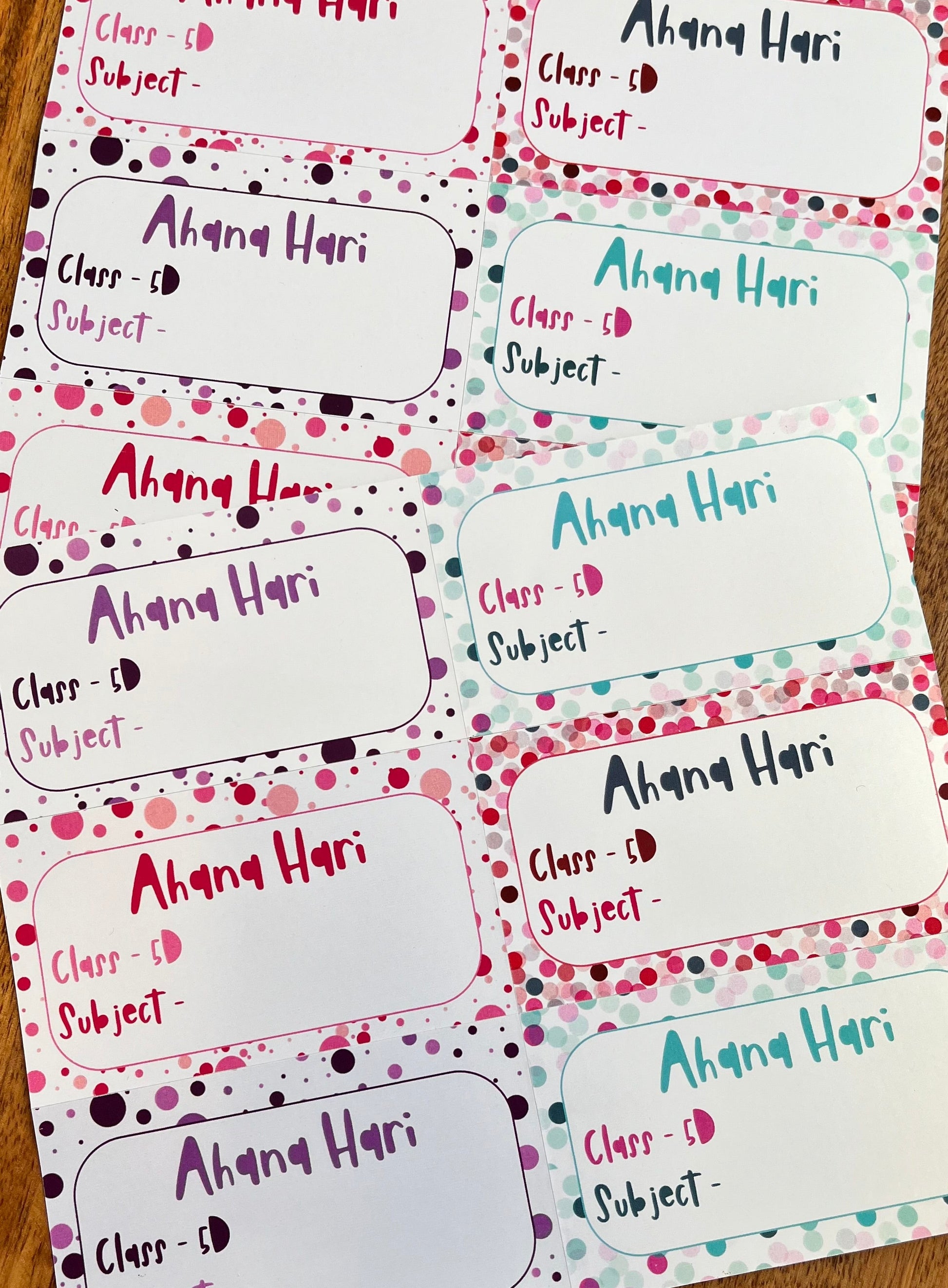 A set of printed class roster labels or name tags laid out on a wooden surface. Each label has a white rounded rectangle body with a decorative border of pink, purple, and teal polka dots on a white background.