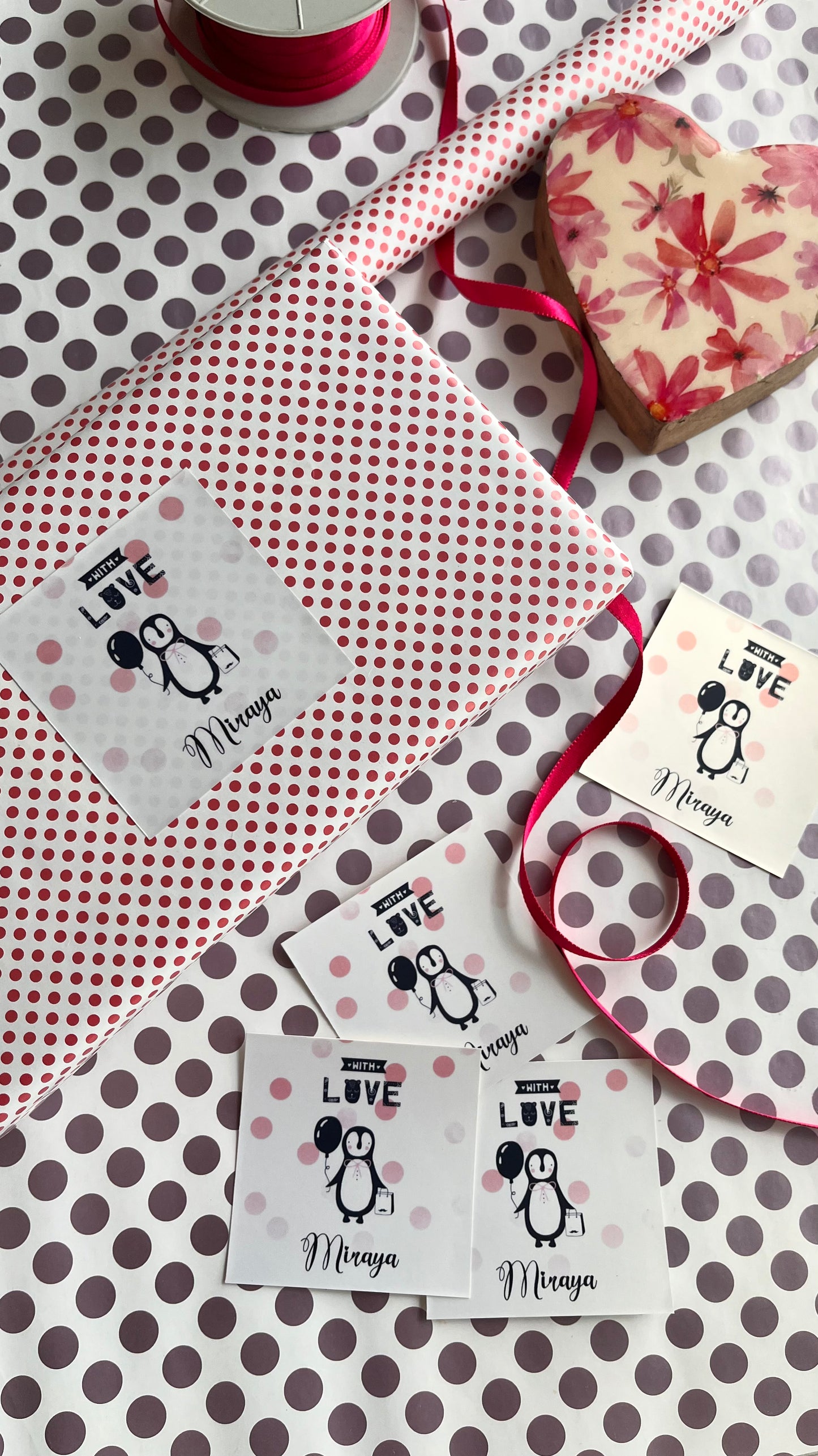 A styled flat-lay of personalized gift wrapping supplies on a white background with brown polka dots. The set includes a roll of red and white polka dot wrapping paper, a heart-shaped box with a pink floral pattern, a spool of pink ribbon, and personalised labels