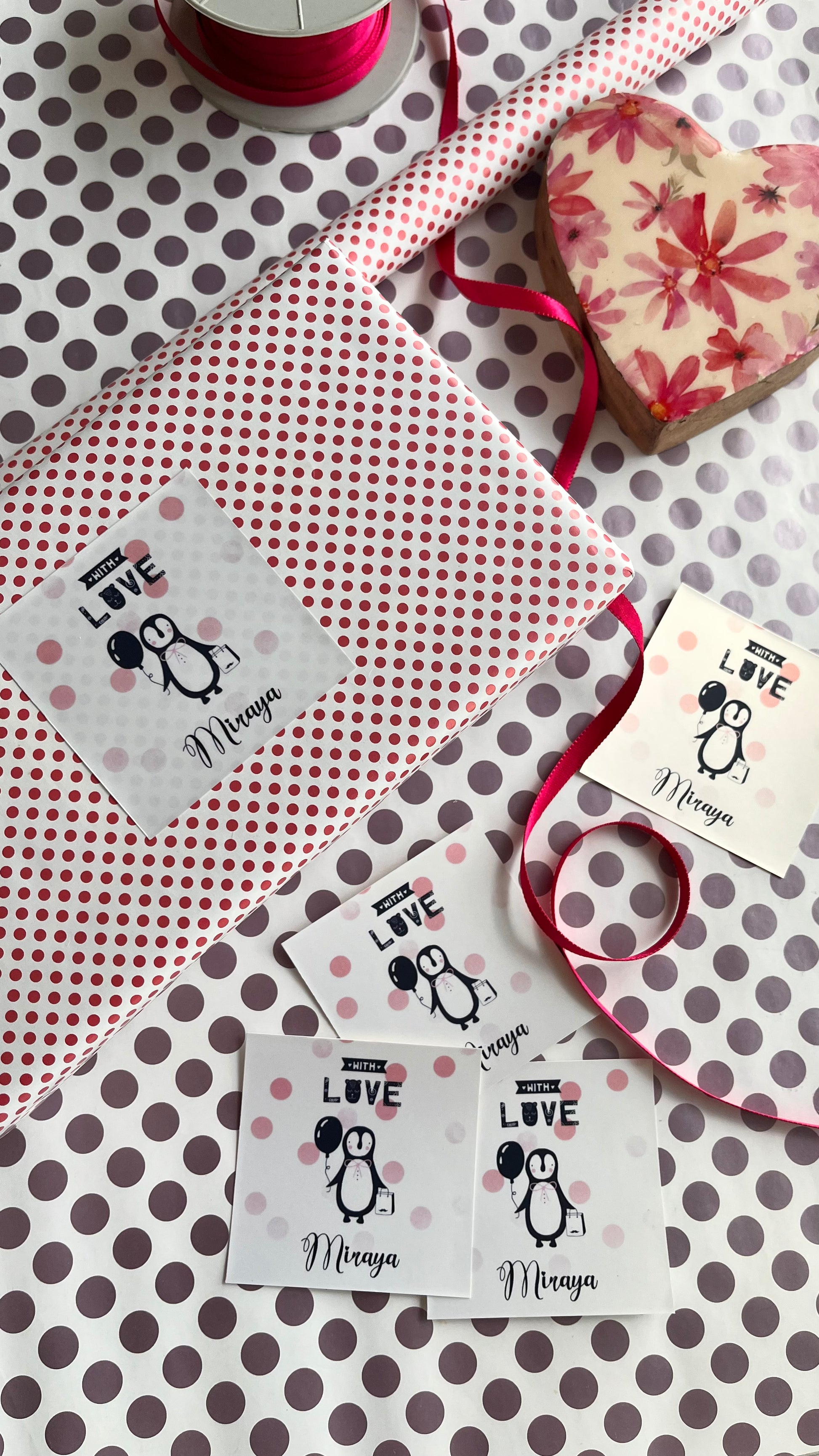 A styled flat-lay of personalized gift wrapping supplies on a white background with brown polka dots. The set includes a roll of red and white polka dot wrapping paper, a heart-shaped box with a pink floral pattern, a spool of pink ribbon, and personalised labels