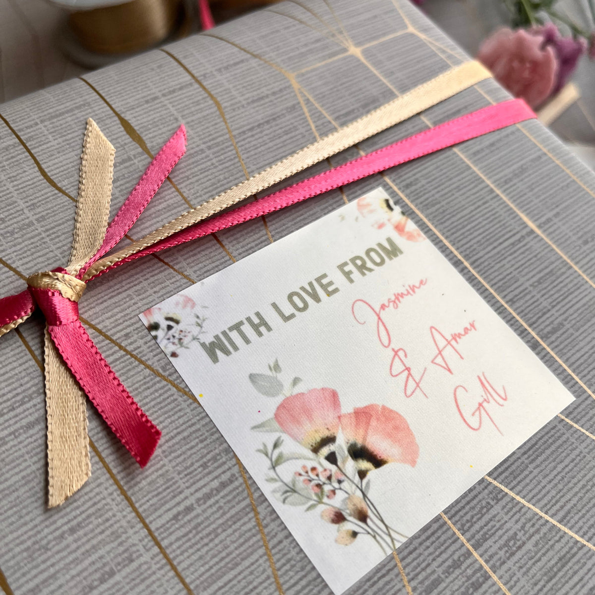 A close-up of a gift box wrapped in gray striped paper with gold lines, tied with a pink and beige ribbon, and featuring a small floral card that reads 'WITH LOVE FROM' and 'My Heart'.
