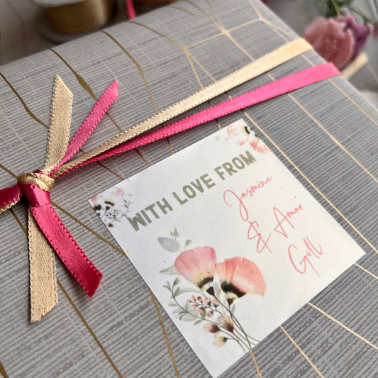 A close-up of a gift box wrapped in gray striped paper with gold lines, tied with a pink and beige ribbon, and featuring a small floral card that reads 'WITH LOVE FROM' and 'My Heart'.