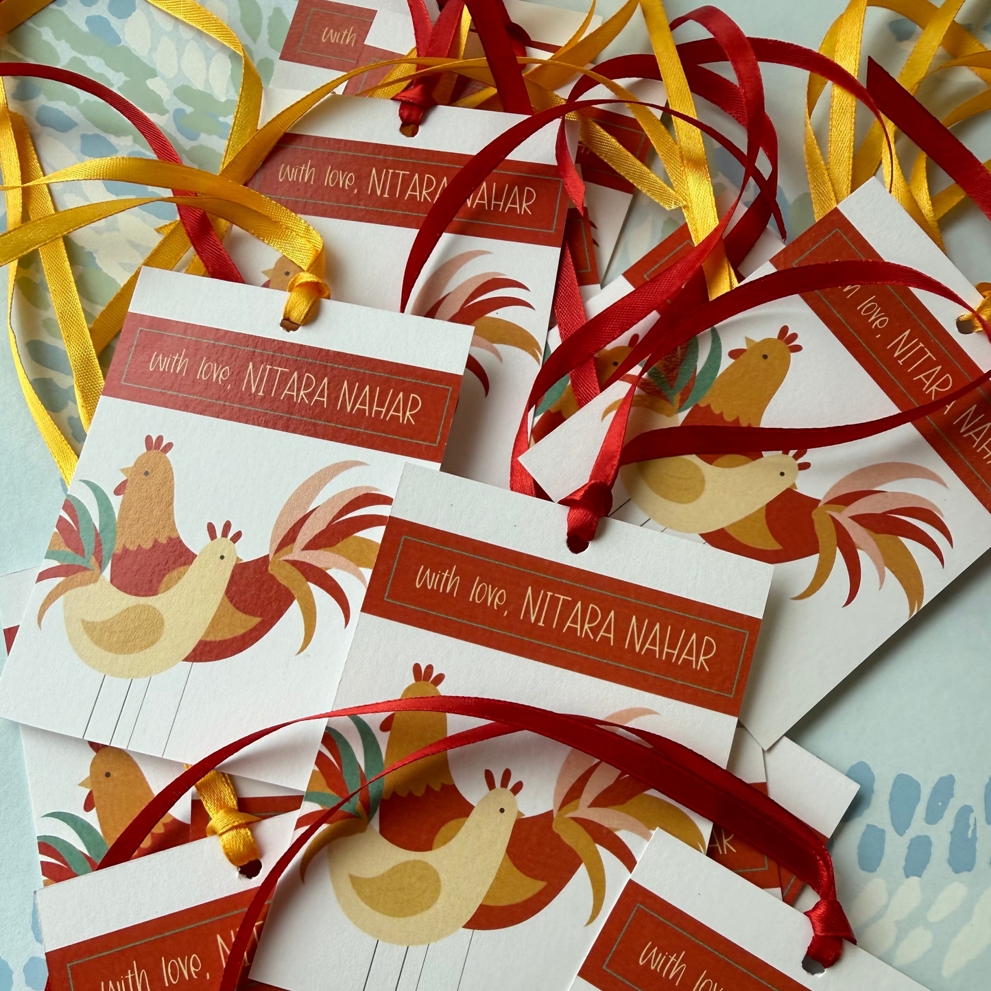 A pile of small printed gift tags or cards with red ribbons, featuring illustrated chickens and the text 'with love, NITARA NAHAR' on a light patterned surface.