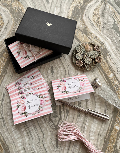 A styled flat-lay of a bridal shower favor kit on a marble-patterned surface. The kit includes a black gift box with a small gold heart, pink and white striped envelopes with floral designs and 'Best Wishes' writing, a silver fountain pen, pink thread, and a decorative butterfly ornament.