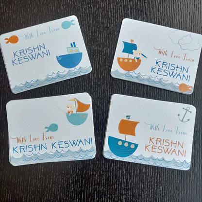 Four personalized baby shower cards arranged on a dark wood background. The cards feature cute cartoon illustrations of ships, boats, fish, and an anchor in shades of blue, orange, and light blue