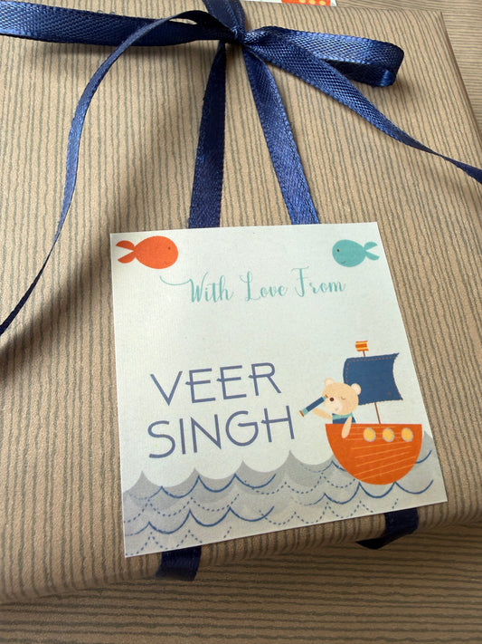 A square gift tag printed with 'With Love From VEER SINGH' and a cartoon child on a small orange boat, attached to a wrapped package with a navy ribbon on a striped kraft paper background.