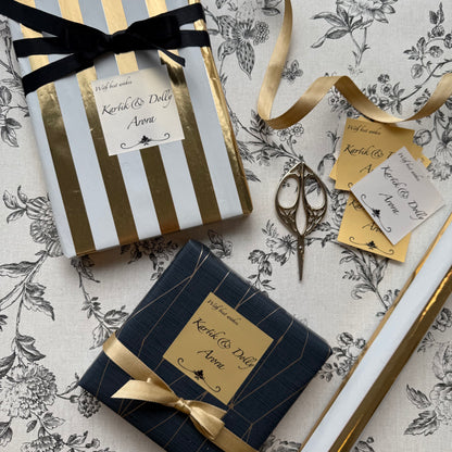 Two wrapped gift boxes on a floral-patterned surface, one with white and gold stripes and a black ribbon, and one with dark navy and gold stripes and a gold ribbon. A gold tag, scissors, and ribbon are arranged around them.