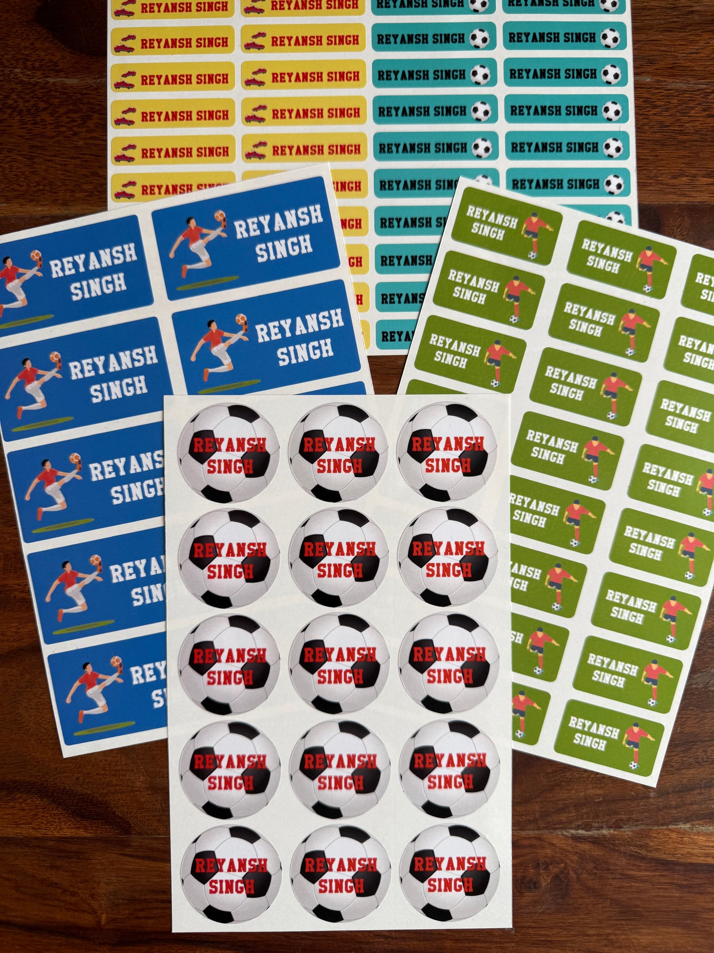 personalised, waterproof school name labels  soccer player graphics, alongside sheets of round football themed stickers and a sheet of rectangular name stickers, all arranged on a dark wood background.
