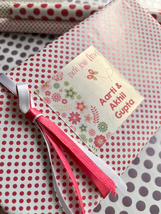 A close-up of a gift-wrapped package featuring pink and white polka dot wrapping paper, a cream floral label that reads 'with love Aarti & Akhil Gupta,' and pink and white ribbons tied around the package.