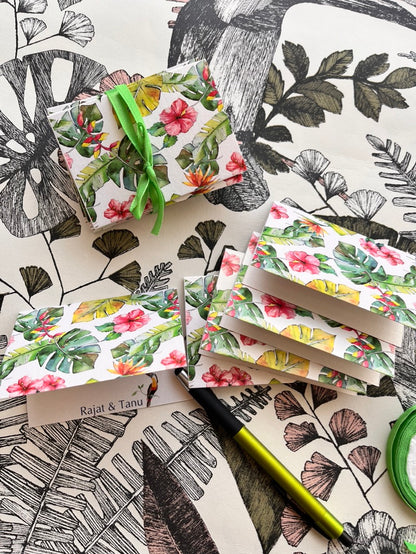 A styled flat-lay of tropical floral gift boxes and matching wrapped packages on a black-and-white botanical patterned surface. The boxes feature pink hibiscus and red flowers with green leaves, tied with bright green ribbon.