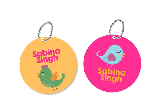Two round metal bag tags with a bird theme. One is pink with a blue bird and the other is green with a yellow bird.