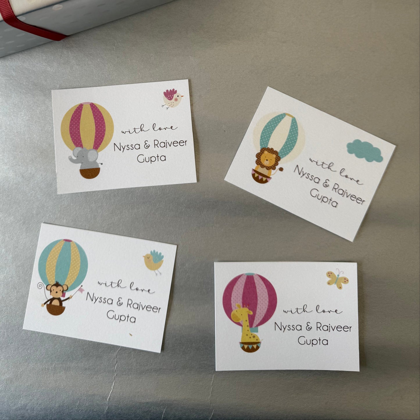 A gift box wrapped alongside four small personalised printed cards featuring colorful hot air balloon illustrations and the personalized text 'with love Nyssa & Raoveer Cupta' on a gray surface.