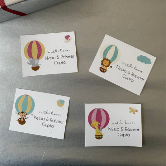 A gift box wrapped alongside four small personalised printed cards featuring colorful hot air balloon illustrations and the personalized text 'with love Nyssa & Raoveer Cupta' on a gray surface.