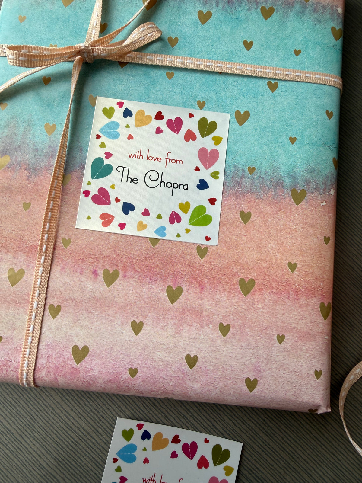 A gift-wrapped package with pastel blue and pink watercolor-style paper, scattered gold heart confetti, a beige ribbon tied in a bow, and a white square card that reads 'with love from The Chakra' surrounded by colorful hearts. A second matching card is partially visible on the right over a gray fabric background.