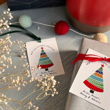 Personalized Christmas tree-shaped gift tags with red and green ribbons, white cardstock, and printed text, displayed on a table with decorative elements.