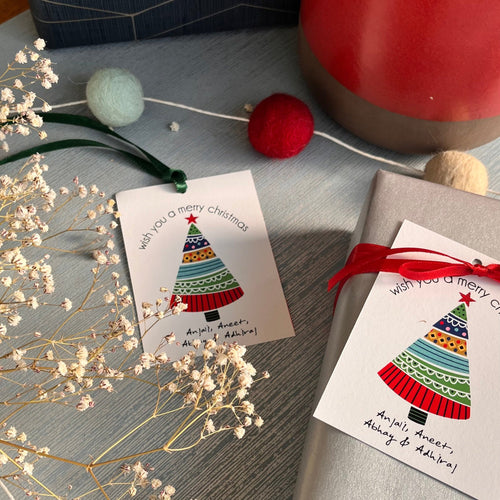 Personalized Christmas tree-shaped gift tags with red and green ribbons, white cardstock, and printed text, displayed on a table with decorative elements.